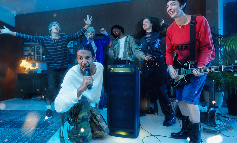 A person singing into the ULTMIC1 next to the TOWER 9 speaker with their friend playing a guitar and the other friends singing in the background