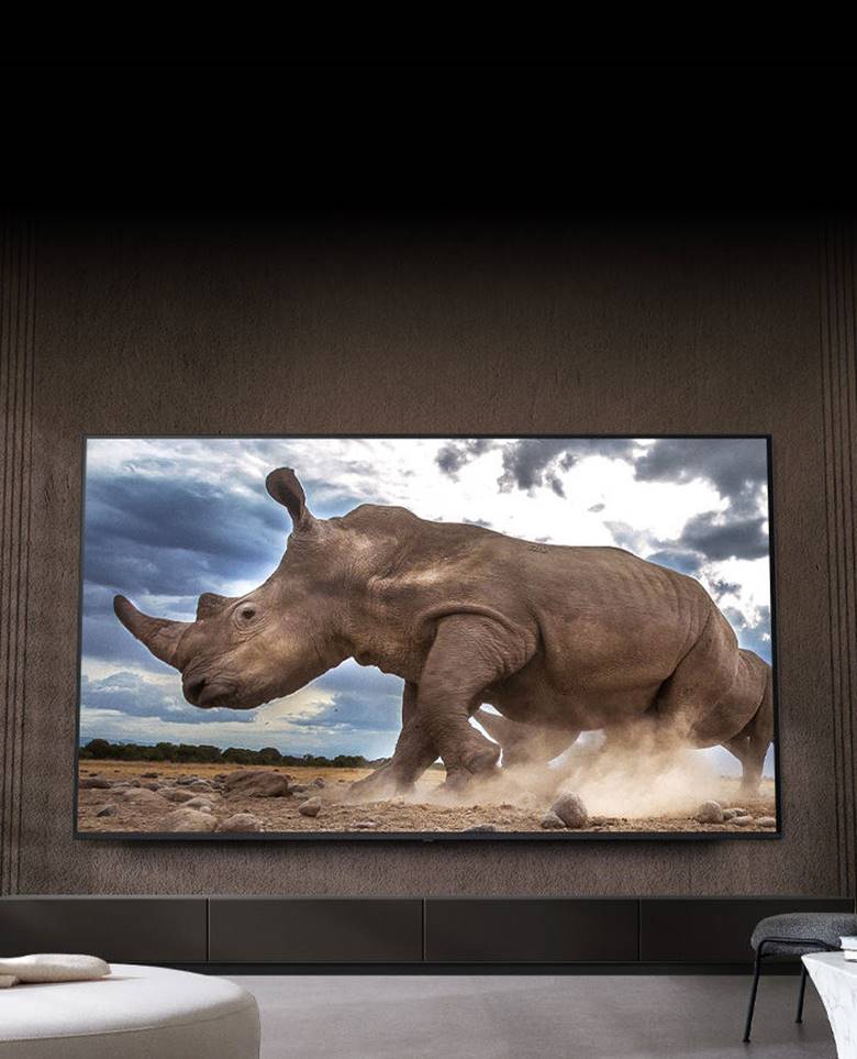 A rhinoceros in a safari setting is shown on an Ultra Big LG TV, mounted on the brown wall of a living room surrounded by cream-colored modular furniture.