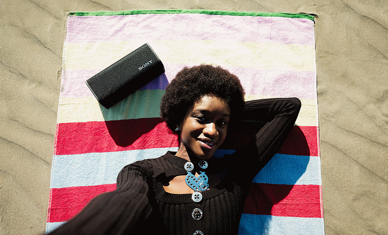 A person laying on their beach towel on the beach with their ULT speaker next to them