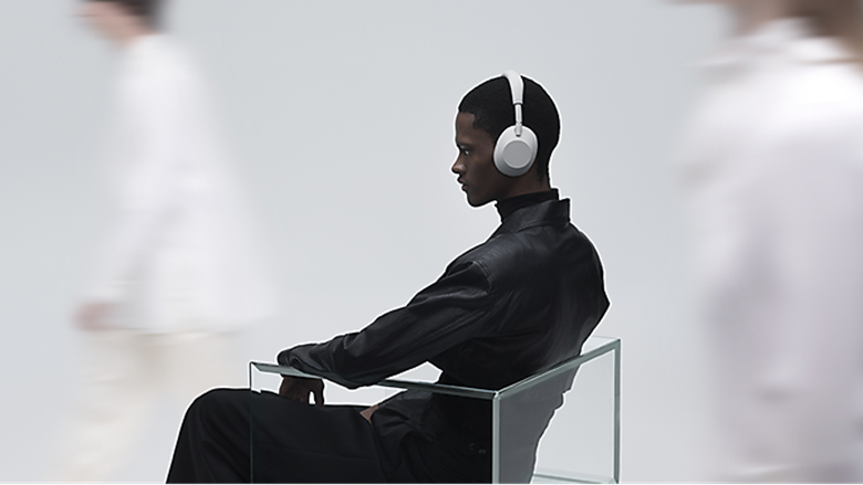 A person sits still wearing a pair of Sony headphones as blurred figures pass by in a minimal white setting.