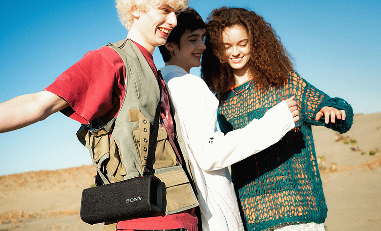 Three people walking on a beach carrying the ULT FIELD 3 speaker using the shoulder strap listening to music