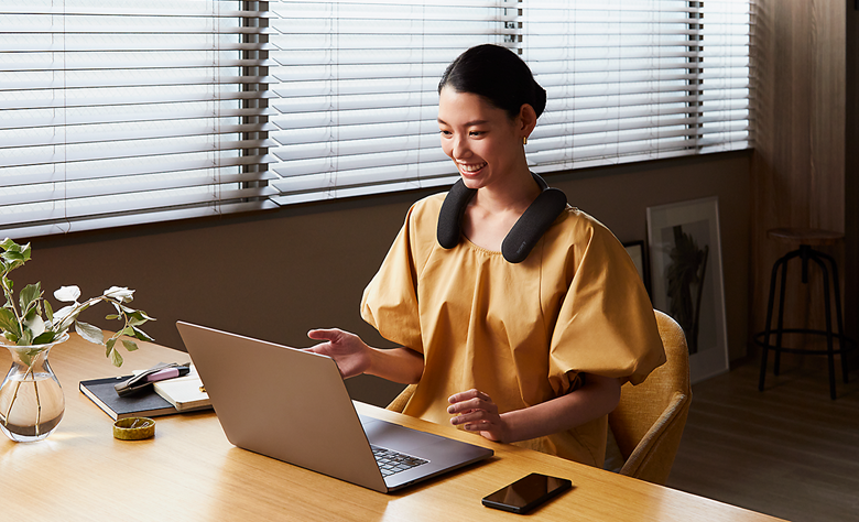 Person sitting at a desk wearing BRAVIA Theatre U on a video call using laptop with notebook and pen on desk to left and smartphone on desk to right 