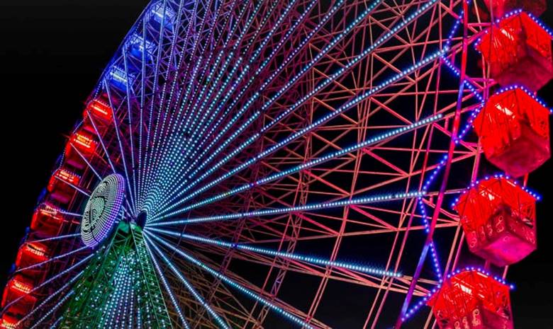 A slightly faded Ferris wheel illuminates at nighttime, with colored sparkles starting from the left side of the screen and moving across it. As the colored sparkles pass, the screen transitions from cloudy to bright, vivid colors.