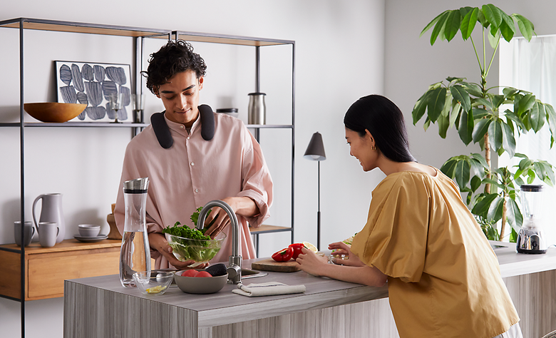 Person wearing BRAVIA Theatre U preparing food in a kitchen with person resting on countertop and ornaments behind on a display unit and large pot plant to right