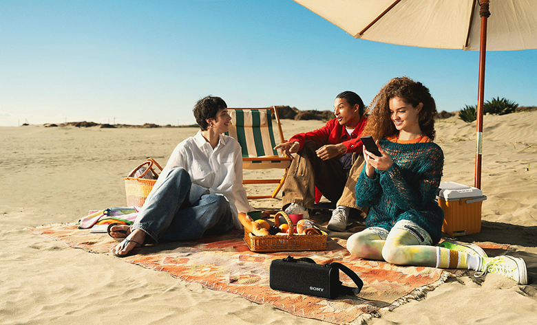 Three people sitting on a beach with the ULT FIELD 3 speaker in front of them, connecting their music through their phone to the speaker