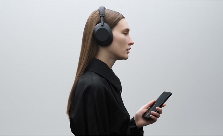 A person in profile view wearing a pair of black WH-1000XM6 headphones while holding a smartphone.