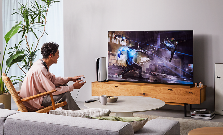 Person sitting on a chair in a living room wearing BRAVIA Theatre U while playing a game on a big screen TV with screenshot of a Spider-Man 2 street scene 