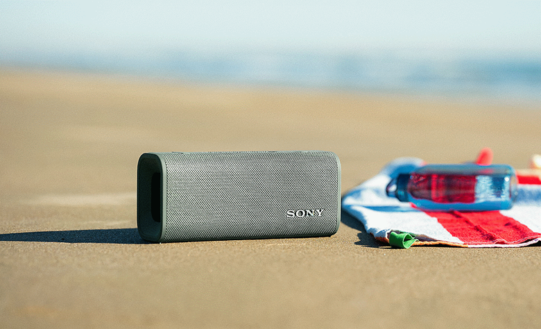The forest green ULT FIELD 3 speaker sitting on the sand on the beach next to a beach towel